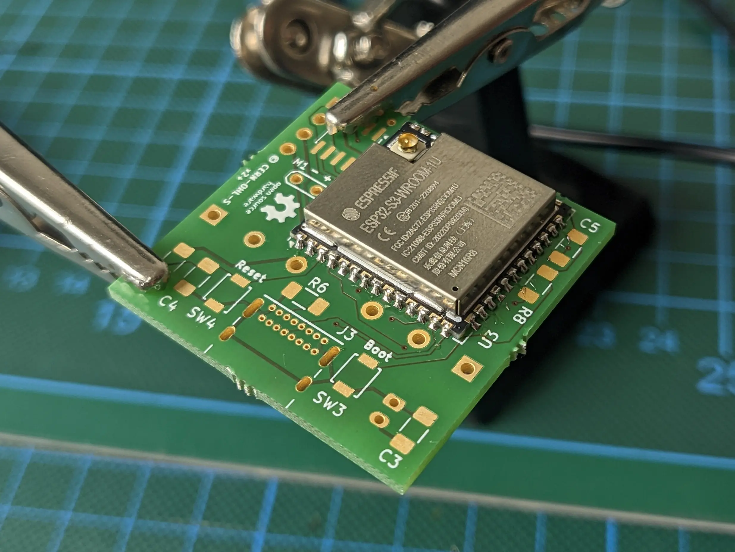Green PCB clibbed onto soldering stand with an ESP32S3 module soldered to it. The other solder pads are still exposed.